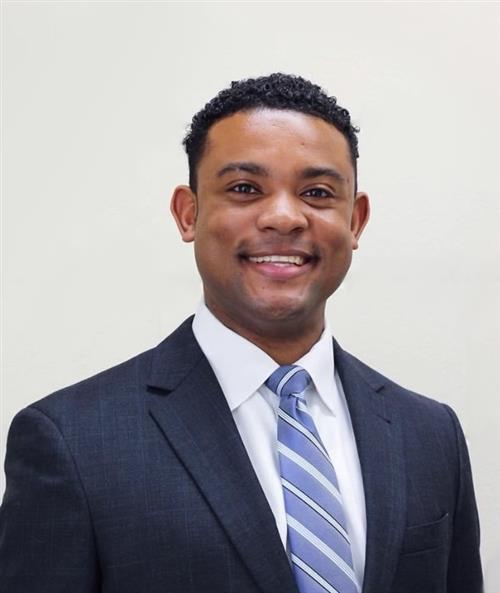 Portrait of a smiling man in a dark suit and blue striped tie against a beige background.